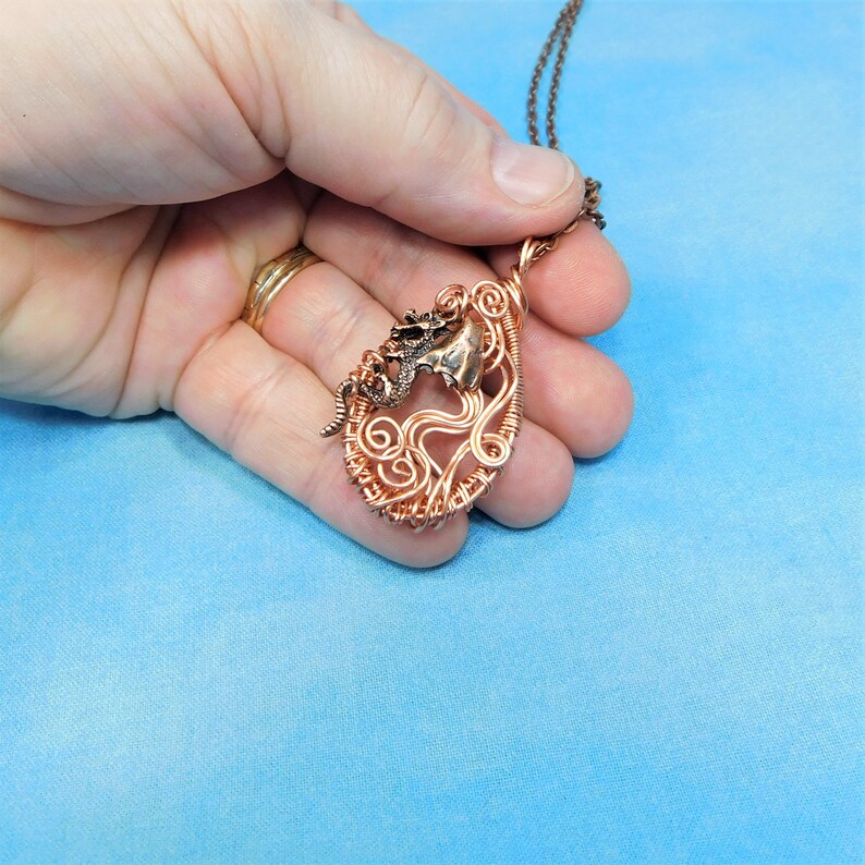 May include: A copper wire wrapped pendant featuring a dragon design. The pendant is in the shape of a heart with a swirl pattern. The dragon is made of a darker metal and is facing to the right.