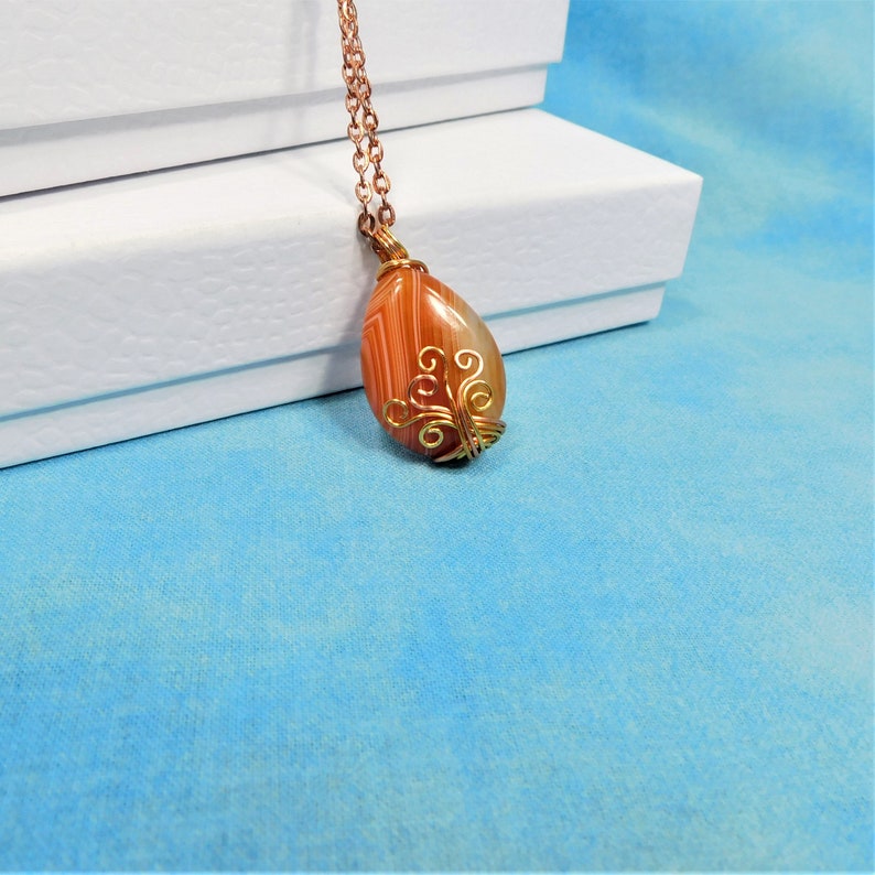 May include: A copper chain necklace with a teardrop-shaped agate pendant. The pendant is wrapped in gold wire with a delicate swirl design. The agate is a mix of orange, brown, and white.