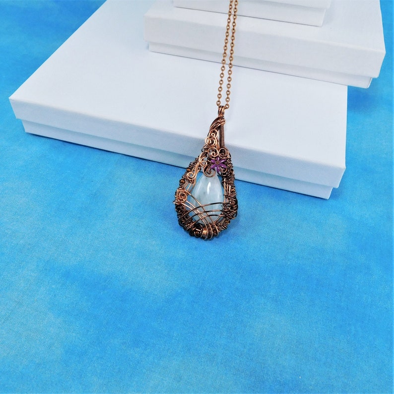 Puede incluir: Un collar con un colgante en forma de l&aacute;grima, compuesto por una piedra preciosa azul claro envuelta en alambre de cobre. El colgante est&aacute; adornado con una peque&ntilde;a flor morada.