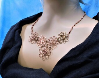 May include: A copper-toned wire necklace featuring a series of interconnected, looped floral-like shapes. The central design consists of multiple swirling wire petals joined together, suspended from a delicate, textured copper-colored chain.
