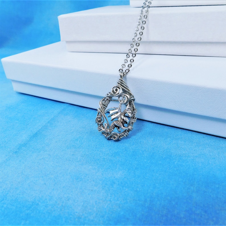 May include: A silver wire wrapped pendant necklace featuring a dragon design. The pendant is oval shaped and hangs from a silver chain.