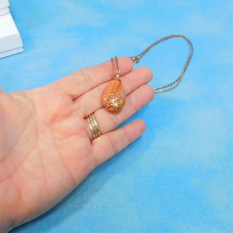 May include: A gold wire wrapped pendant with a teardrop shaped agate stone. The stone is a mix of orange, brown, and white colors. The pendant is hanging from a silver chain.