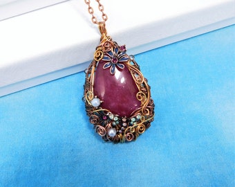 Puede incluir: Un collar con un colgante en forma de lágrima, que contiene una piedra preciosa rosa rodeada de alambre de cobre y pequeñas cuentas. El colgante está adornado con una flor azul con un centro rosa.