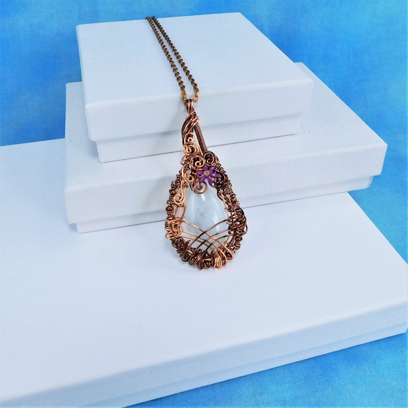 Puede incluir: Un colgante de piedra blanca en forma de l&aacute;grima con un envoltorio de alambre de cobre y un peque&ntilde;o acento de flor morada. El colgante cuelga de una cadena de cobre.