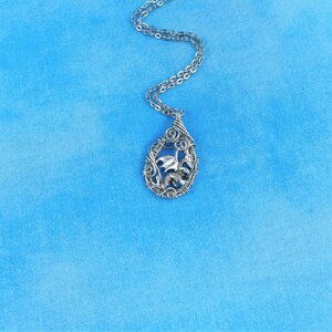 May include: A silver necklace with a dragon pendant. The dragon is made of wire and is in a flying pose. The necklace is on a blue background.