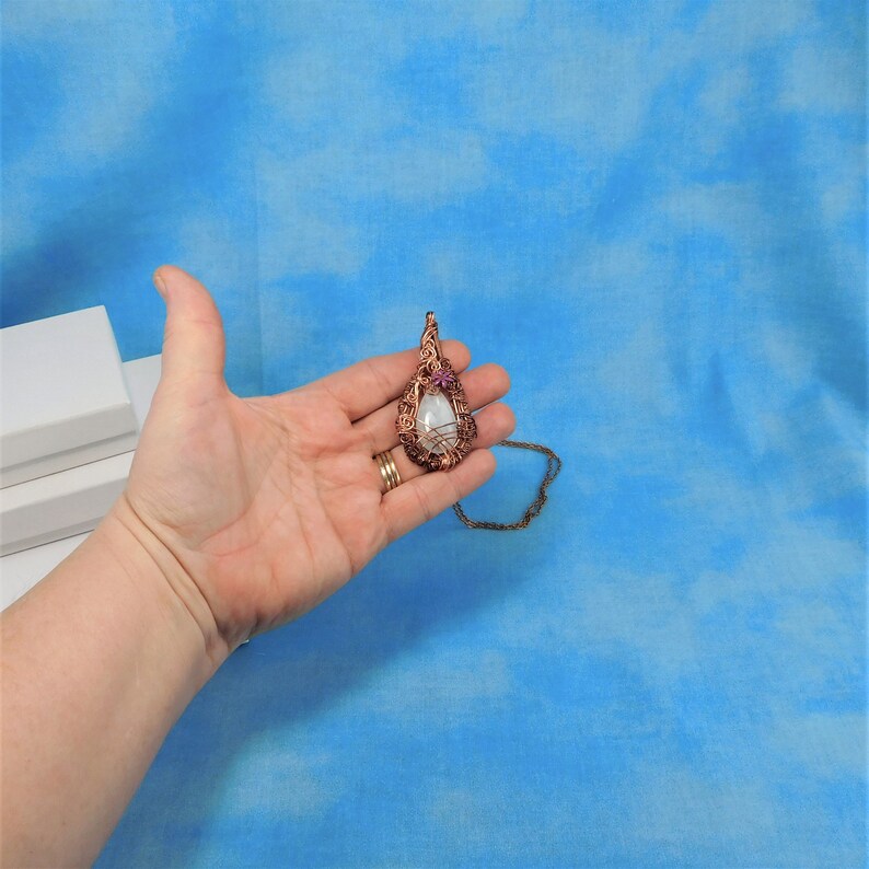 Puede incluir: Un collar con un colgante en forma de l&aacute;grima que contiene una piedra blanca envuelta en alambre de cobre. El colgante est&aacute; adornado con una peque&ntilde;a flor morada. El collar est&aacute; en una cadena marr&oacute;n.