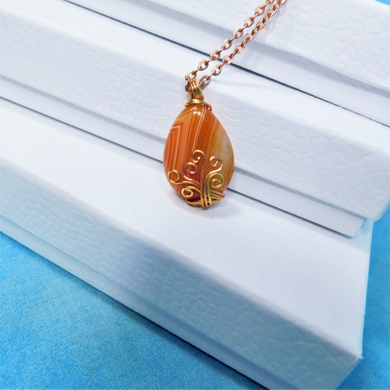 May include: A gold wire wrapped pendant featuring a teardrop-shaped agate stone with orange and white stripes. The pendant is hanging from a gold chain.