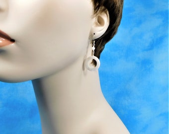 May include: A drop earring featuring a small white bead connected to a larger, smooth white circular ring. The earring is suspended from a silver-toned metal hook, creating a simple and clean design that hangs vertically from the earlobe.