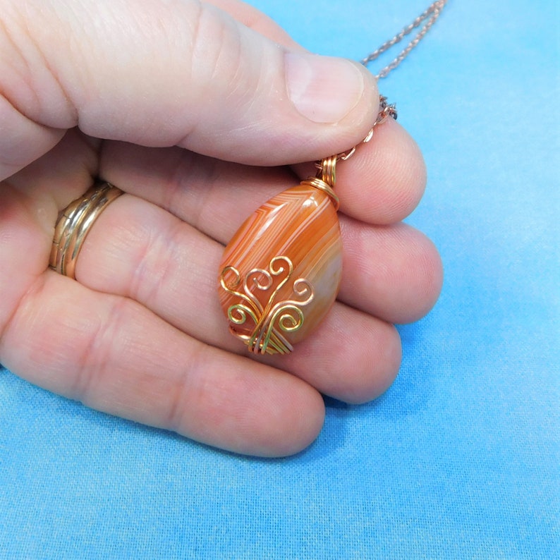 May include: A teardrop-shaped carnelian agate pendant with a gold wire wrapped design. The pendant is hanging from a gold chain.