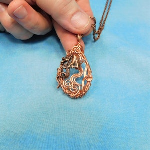 May include: A copper wire necklace with a pendant featuring a dragon design. The pendant is made of copper wire and has a teardrop shape. The dragon is curled up in the pendant and has a detailed design.