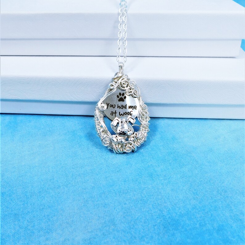 May include: Silver necklace with a teardrop-shaped pendant. The pendant is made of wire wrapped around a silver disc with the text "You had me at woof" and a paw print.