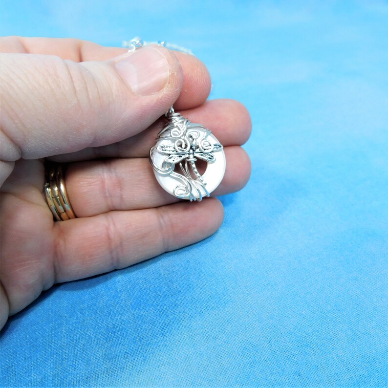 May include: A silver wire-wrapped pendant featuring a white round stone with a dragonfly design.