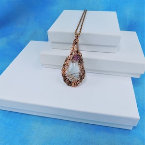 Puede incluir: Un collar con un colgante en forma de l&aacute;grima que contiene una piedra de vidrio transparente enmarcada en alambre de cobre. El marco est&aacute; decorado con una peque&ntilde;a flor morada.
