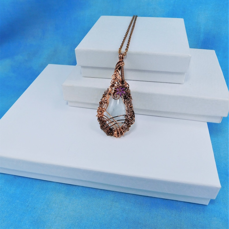Puede incluir: Un collar con un colgante en forma de l&aacute;grima que contiene una piedra de vidrio transparente enmarcada en alambre de cobre. El marco est&aacute; decorado con una peque&ntilde;a flor morada.