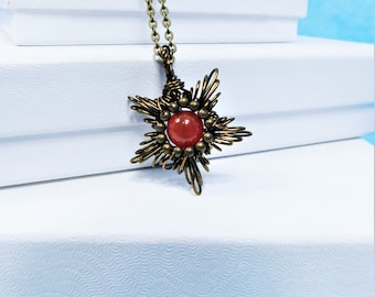 May include: A necklace featuring a star-shaped pendant crafted from dark bronze-toned wire. The center of the pendant contains a smooth, round, reddish-orange bead surrounded by smaller bronze-toned metal spheres, suspended on a matching chain.
