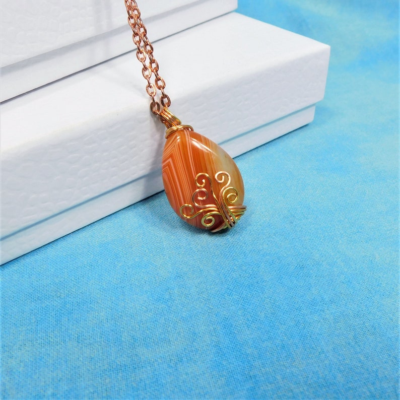 May include: A teardrop-shaped carnelian agate pendant with a gold wire wrap design. The pendant is hanging from a copper chain.