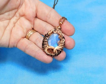 May include: A wire-wrapped pendant necklace featuring a central oval-shaped iridescent blue moonstone cabochon. The stone is encased in intricate, dark-toned copper wire coils and swirls, suspended from a matching dark copper metal chain.