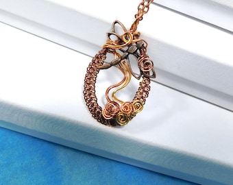Puede incluir: Un colgante de alambre envuelto en cobre y oro con un diseño de libélula. El colgante tiene forma ovalada y cuelga de una cadena de cobre.