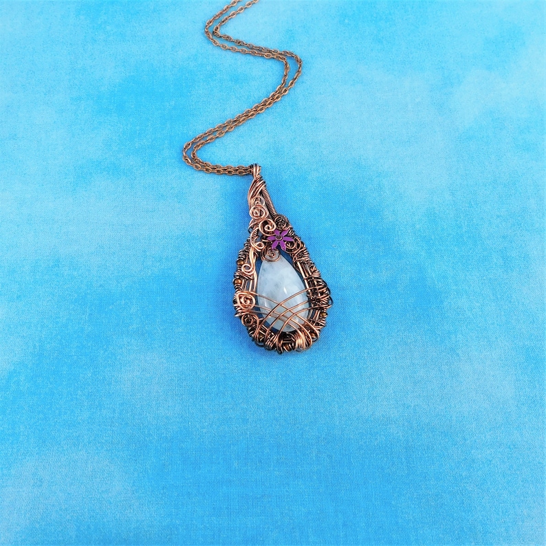Puede incluir: Un collar con un colgante en forma de l&aacute;grima, con una piedra blanca envuelta en alambre de cobre. El colgante tiene un peque&ntilde;o adorno de flor morada.