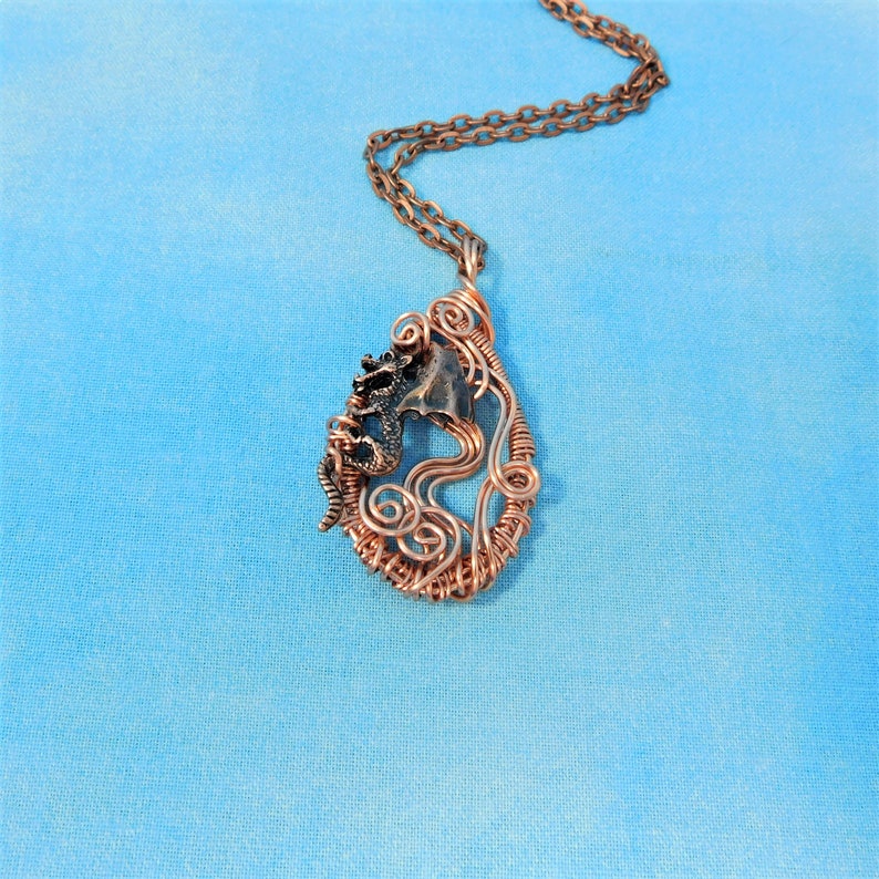 May include: A copper wire wrapped pendant necklace featuring a small dragon figurine. The pendant is shaped like a teardrop and has a swirling wire design.