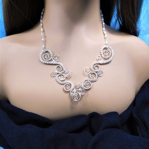 May include: A silver-toned wire necklace with a swirling, scroll-like design. The necklace features multiple spiral elements connected to a beaded chain. The design is symmetrical and sits on a mannequin.