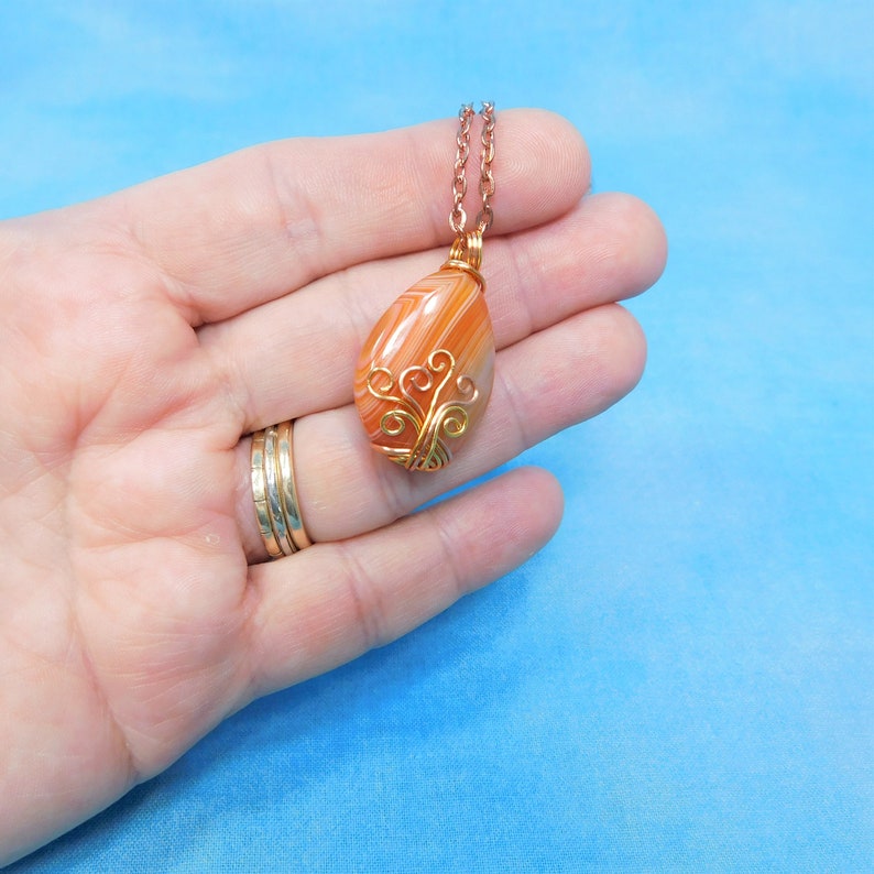 May include: A teardrop-shaped pendant necklace with a wire-wrapped agate stone. The stone is orange and white with a swirling pattern. The wire is gold-colored and has a delicate design.
