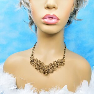 May include: A bronze-toned metal necklace featuring a series of interconnected wire spirals arranged in a V-shaped pattern. The chain consists of small, round metallic beads that connect to the central spiral design, creating a textured look.
