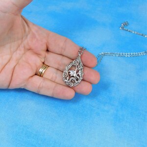 May include: A silver wire-wrapped pendant featuring a dragon design hangs from a silver chain. The pendant is held in a hand against a blue background.