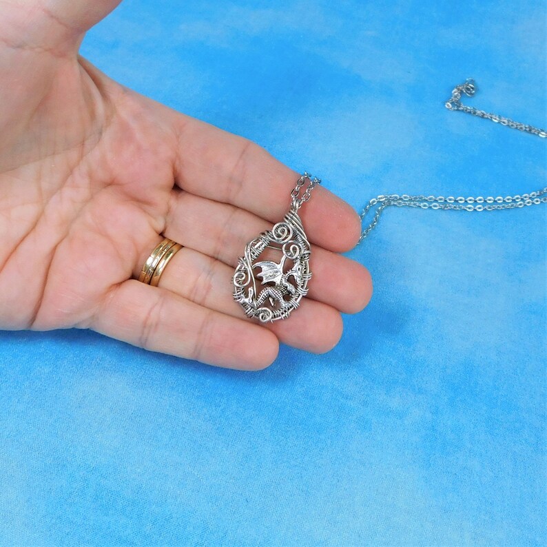 May include: A silver wire-wrapped pendant featuring a dragon design hangs from a silver chain. The pendant is held in a hand against a blue background.