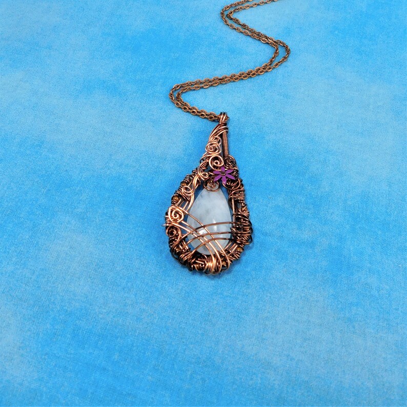 Puede incluir: Un collar con un colgante de alambre de cobre envuelto alrededor de una piedra preciosa blanca con un acento de flor morada.