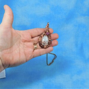 Puede incluir: Un collar con un colgante en forma de l&aacute;grima, con una piedra blanca envuelta en alambre de cobre. El colgante tiene un peque&ntilde;o acento de flor rosa. El collar est&aacute; en una cadena de cobre.