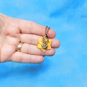 Puede incluir: Un collar de oro con un colgante de flor amarilla con un diseño de abeja. El colgante está hecho de alambre y tiene un diseño delicado e intrincado.