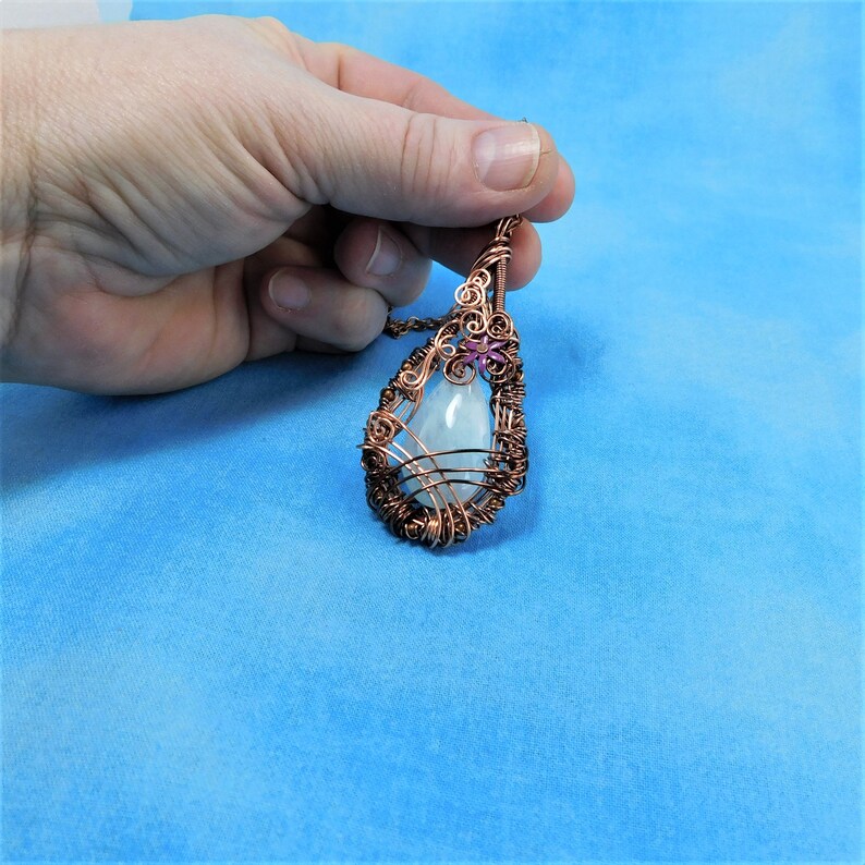 Puede incluir: Un colgante en forma de l&aacute;grima con una piedra preciosa blanca envuelta en alambre de cobre. Una peque&ntilde;a flor rosa est&aacute; unida a la parte superior del colgante.