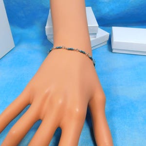 May include: A delicate bracelet featuring small, cylindrical beads in shades of blue and black, strung together with silver-colored links. The bracelet is displayed on a mannequin hand against a blue background, with white boxes in the background.