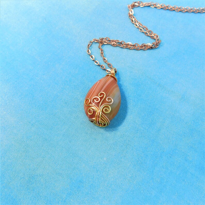 May include: A gold wire wrapped pendant featuring a teardrop-shaped agate stone with brown and white stripes. The pendant is attached to a gold chain.