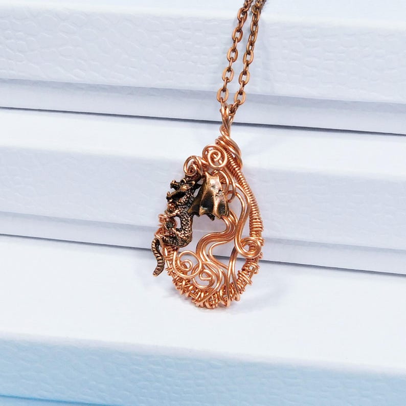 May include: A copper wire wrapped pendant necklace features a small dragon figurine. The pendant is shaped like a teardrop and has a delicate, intricate design.