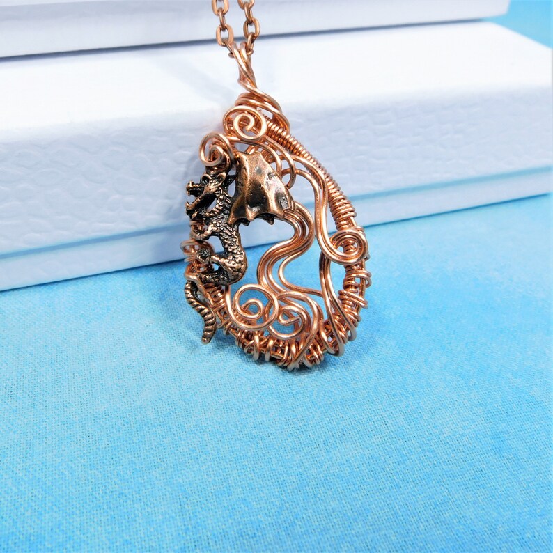 May include: A copper wire wrapped pendant featuring a small dragon figurine. The pendant is shaped like a teardrop and has a delicate, intricate design.