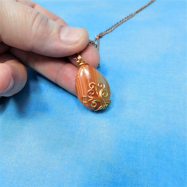 May include: A copper chain necklace with a pendant featuring a brown and orange agate stone wrapped in gold wire with a decorative design.