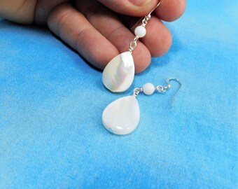 May include: A pair of drop earrings featuring iridescent white teardrop-shaped shell pendants suspended from silver-toned hooks. Each earring includes a small, round white bead accent connected by silver-toned wire links to the main shell drop.