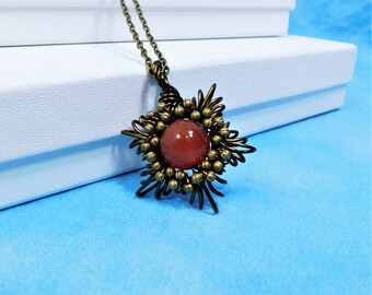 May include: A pendant necklace featuring a round, smooth, reddish-orange stone at the center. The stone is surrounded by a dark bronze-toned wire-wrapped frame with small metallic beads, suspended from a matching dark bronze-toned metal chain.