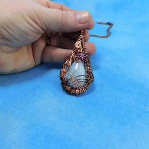 Puede incluir: Un colgante en forma de l&aacute;grima con una piedra preciosa blanca envuelta en alambre de cobre. El colgante presenta una peque&ntilde;a flor morada con un tallo de alambre de cobre.