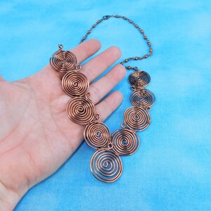 K&ouml;nnte beinhalten: Eine Halskette aus Kupferdraht mit einem kaskadierenden Design aus mehreren Spiralkreisen. Die Halskette liegt auf einer Hand vor blauem Hintergrund.