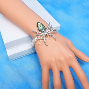 May include: Silver-toned spider cuff bracelet with an abalone shell center and crystal accents. The spider's legs extend outward, and the bracelet is worn on a wrist. The bracelet is displayed on a mannequin hand against a blue background.