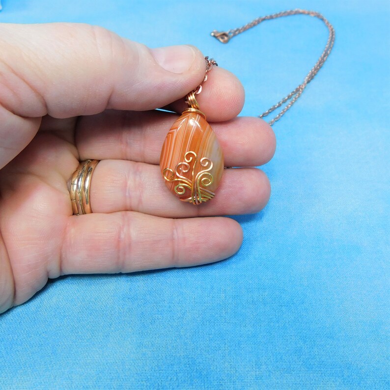 May include: A copper wire wrapped agate pendant with a tree design. The pendant is on a copper chain and is being held in a hand.