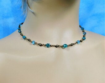 May include: A delicate choker necklace featuring a series of small, faceted turquoise blue crystal beads alternating with textured, antique bronze-toned metal spacer beads, all connected by thin, dark metal wire links in a simple, repeating pattern.