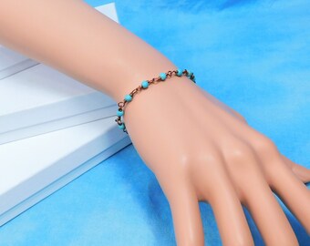 May include: A delicate bracelet featuring small, round, bright turquoise beads spaced evenly along a thin, dark copper-toned metal chain. The chain consists of interconnected circular links, creating a simple and lightweight design that rests on the wrist.