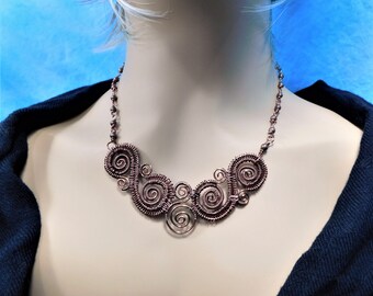 May include: A copper wire necklace featuring a series of interconnected spiral motifs. The central design consists of multiple coiled wire circles of varying sizes, linked together to form a wide, decorative collar piece with a beaded chain.