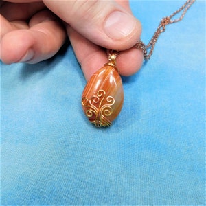 May include: A teardrop-shaped agate pendant with orange and white stripes, wrapped in gold wire with a delicate, swirling design.