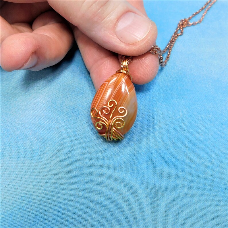May include: A teardrop-shaped agate pendant with orange and white stripes, wrapped in gold wire with a delicate, swirling design.