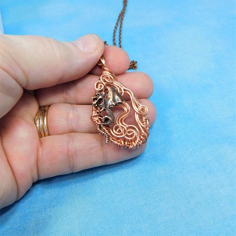 May include: A copper wire wrapped pendant necklace featuring a small dragon figurine. The dragon is curled up in a wire frame that resembles a cloud or wave.